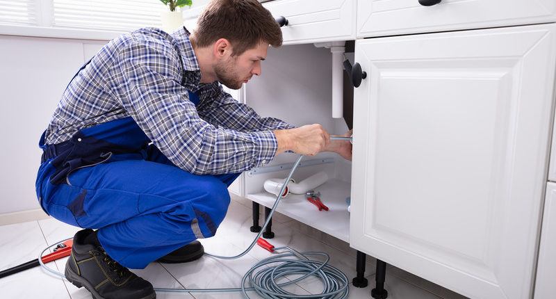 Young Male Plumber Cleaning Clogged Sink Pipe In Kitchen